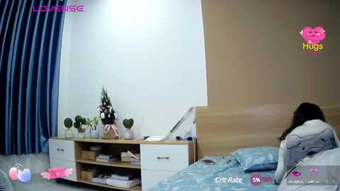 Media: Video of a woman sleeping in a minimalist bedroom with white walls, wooden headboard, blue bedspread, and potted plants on a wooden nightstand.