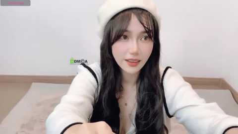 Media: Video of a young Asian woman with long black hair, fair skin, and brown eyes, wearing a white beanie, white jacket, and black top, smiling, sitting on a beige rug.