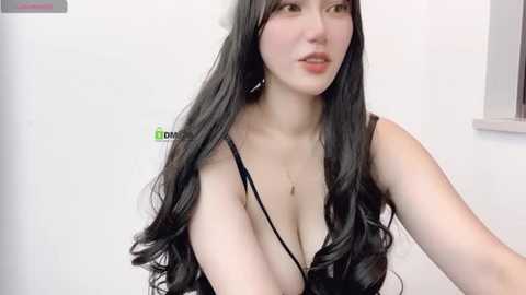 Media: A video of a young Asian woman with long, black wavy hair, wearing a black spaghetti strap top that reveals cleavage, in a white kitchen setting.