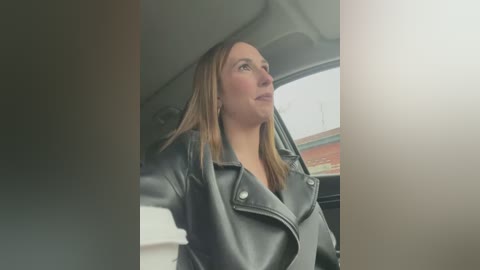 Media: Video of a blonde woman with light skin, wearing a black leather jacket, looking out of a car window at a cloudy sky.