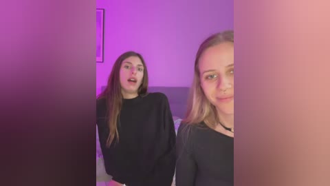 Media: Video of two young women with light skin, long hair, and black clothing in a dimly lit room with purple lighting, smiling and looking at the camera.