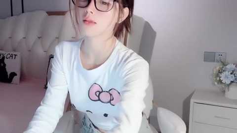 Media: Video of a young Asian woman with glasses, wearing a white T-shirt with a Hello Kitty design, sitting on a bed with a tufted headboard.