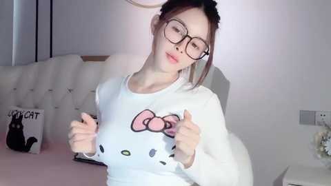 Media: Video of a young Asian woman with fair skin, glasses, and brown hair, wearing a white shirt with a Hello Kitty design. She stands in a modern, softly lit bedroom with white walls and a plush bed.