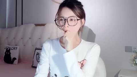 Media: A video of a young Asian woman with glasses, wearing a white top, sitting on a bed with a tufted headboard, holding a phone.