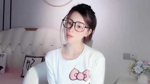 Media: A video of a fair-skinned, slender woman with dark hair, wearing black-rimmed glasses, a white shirt with a pink bow graphic, and a pendant necklace, seated in a modern, minimalist bedroom with a tufted white headboard, floral arrangement, and a lamp.