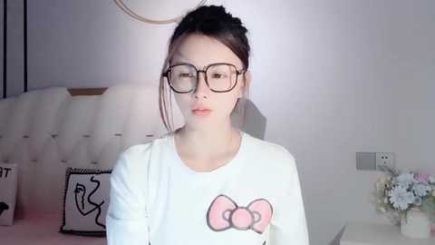 Media: Video of an Asian woman with glasses and dark hair tied back, wearing a white shirt with a pink bow graphic, standing in a minimalist bedroom with a tufted headboard, floral decor, and a beige wall.