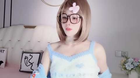 Media: Video of a young Asian woman with straight, shoulder-length blonde hair, wearing glasses, light blue off-shoulder sweater, and a pink heart sticker on her forehead. Background features a white tufted headboard, black and white artwork, and floral arrangement.