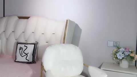 Media: Video of a chic, minimalist bedroom with a white tufted headboard, a black-and-white checkered pillow, and a small white table holding a vase of light pink and blue flowers.