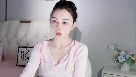 Media: Video of a fair-skinned woman with dark hair in a bun, wearing a pink top, sitting on a white tufted bed in a minimalist room with a floral arrangement on a nightstand.