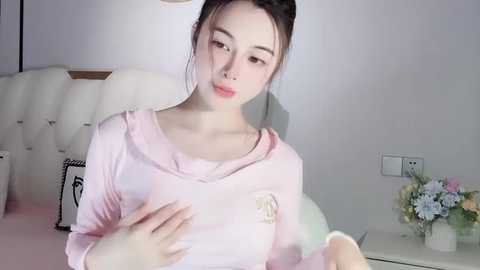 Media: A video of a young Asian woman with fair skin, wearing a pink hoodie, sitting in a modern, minimalist bedroom with white walls and a floral arrangement on a side table.