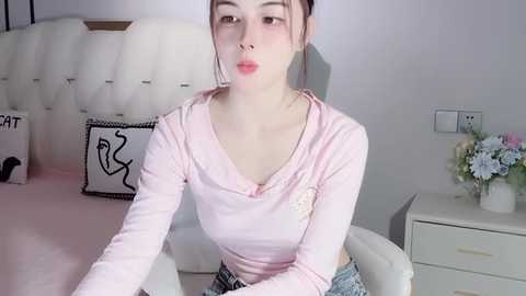 Media: Video of an Asian woman with light skin, brown hair tied back, wearing a light pink long-sleeved top and blue denim shorts, sitting on a white chair in a minimalist bedroom.