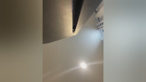 Media: Video of a modern, minimalist room with beige walls and a large, sleek, metallic ceiling fan. The room is illuminated by a single, bright light source, casting a soft glow. The ceiling fan's blades are partially visible, adding a touch of contemporary style.