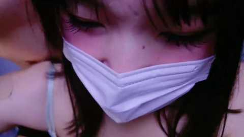 Media: A close-up video of an Asian woman with long dark hair, wearing a white surgical mask, her eyes closed, appearing to be in deep thought or sleep.