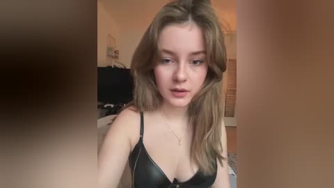 Media: A video of a young Caucasian woman with long, straight brown hair styled with a side part. She has a fair complexion, light makeup, and is wearing a black bra. The background shows a cluttered room with a TV and various objects.