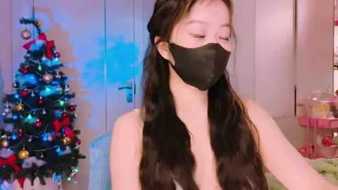 Media: Video of a young Asian woman with long black hair, wearing a black mask, standing in a brightly lit room with a decorated Christmas tree and presents in the background.