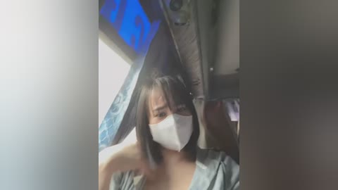 Media: Video of an Asian woman with straight, shoulder-length black hair, wearing a white face mask and a green shirt, seated on a train.