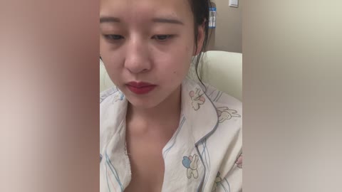 Media: Video of a young East Asian woman with fair skin and black hair in a bun, wearing a white floral-patterned robe, looking pensive in a beige room.