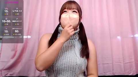 Media: Video of an Asian woman with long brown hair in pigtails, wearing a grey turtleneck sweater, covering her mouth with her hand, against a pink curtain background.