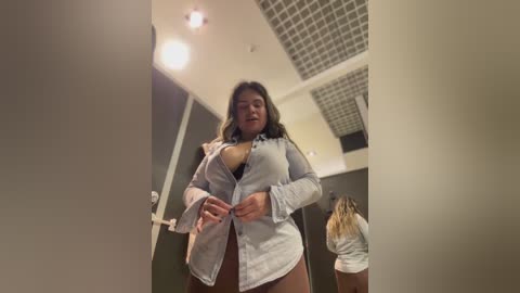 Media: Video of a woman in a dimly lit restroom, wearing a partially unbuttoned white shirt, exposing her chest, with a shocked expression. Another woman, with long blonde hair, stands in the background.