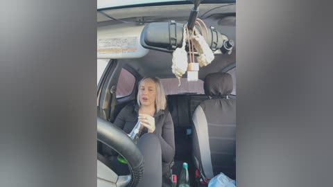 Media: Video of a blonde woman in a black top and jeans driving a car, with a dashboard full of personal items, including a phone and a water bottle.