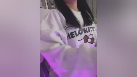 Media: A video of a person with straight, black hair wearing a white sweatshirt with \"RELAXKIT\" and cartoon glasses. The background includes a blurred, white wall with framed pictures.