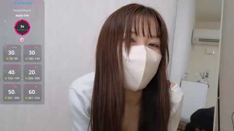 Media: A video of a woman with long brown hair wearing a white surgical mask, standing in a bathroom with a white wall, air conditioner, and a toilet.