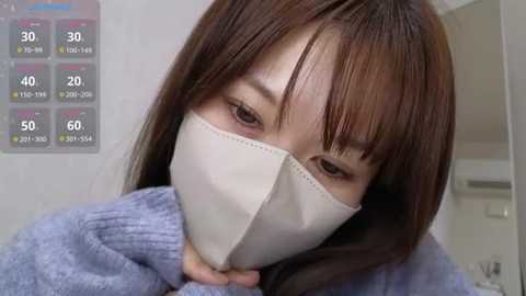 Video of an East Asian woman with straight brown hair, wearing a white face mask, gray sweater, and brown eyes. Background shows a digital temperature gauge with readings.