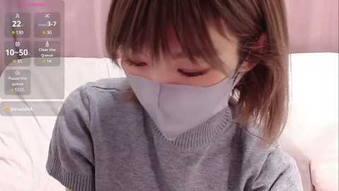 Media: Video of a young East Asian woman with straight brown hair in a ponytail, wearing a gray sweater and a light blue face mask, sitting indoors on a bed with pink curtains in the background.