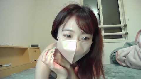 Media: Video of an Asian woman with long, dark hair and fair skin wearing a surgical mask, lying on a bed with a beige sheet in a simple, brightly lit room with wooden furniture.