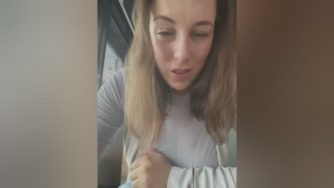 Media: Video of a Caucasian woman with light skin, long blonde hair, wearing a light gray sweater, looking down with a concerned expression, inside a car with dark upholstery.