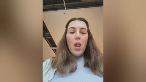 Media: Video of a young woman with long, light brown hair, wearing a white turtleneck, standing in a beige room with visible ceiling beams, looking up with her mouth open, possibly mid-scream.