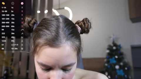 Media: Video of a woman with light skin and brown hair styled in two small buns, wearing a strapless top, indoors with blurred background featuring a Christmas tree and modern decor.