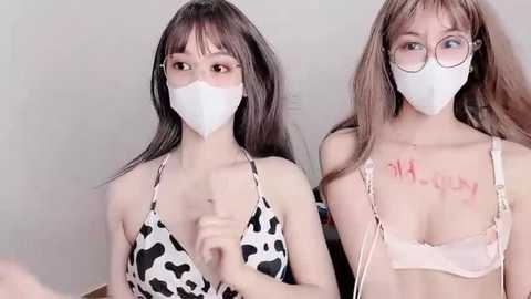 Media: Video of two young Asian women with long hair, wearing cow-patterned tops and face masks, standing indoors with a neutral background.