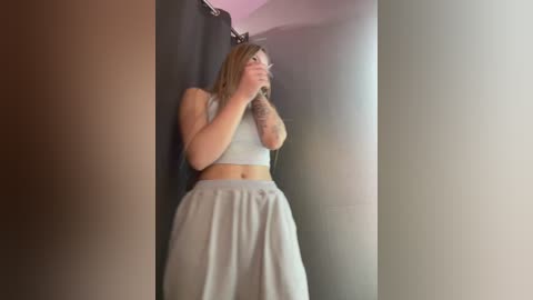 Media: A video of a slender woman with light skin, wearing a light gray crop top and high-waisted pants, stands in a dimly lit, narrow hallway, her hands covering her face.