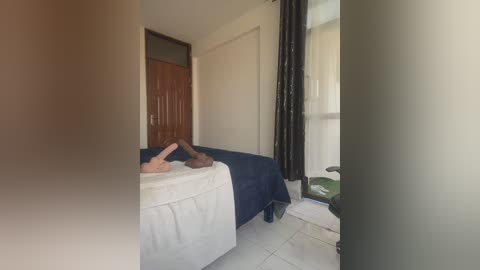 Media: A video of a small, sparsely furnished bedroom with a blue bed, white sheet, and brown wooden door. A person's legs are visible, wearing brown socks. The room has white tiled floor and a window with black curtains.