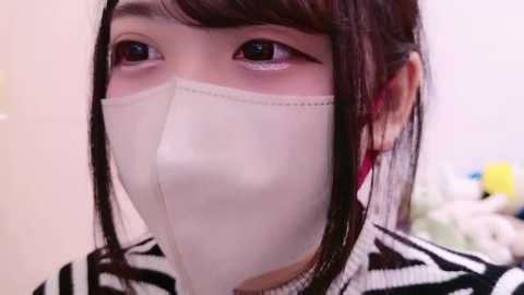 Media: Video of an Asian woman with fair skin, wearing a white surgical mask, black and white striped top, and straight brown hair. Background is blurred, showing a soft, neutral indoor setting.