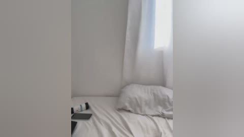 Media: Video of a minimalist, white-walled room with a single bed, neatly made with white sheets, a pillow, and a small nightstand beside it. A window with sheer white curtains allows soft, diffused light to enter.