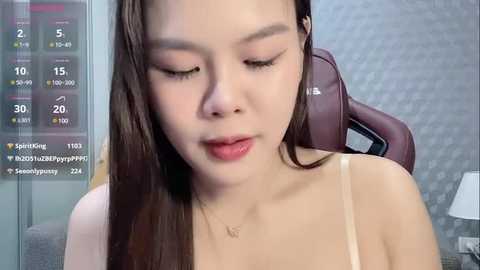 Media: Video of a young Asian woman with long, straight brown hair, wearing a beige spaghetti strap top, sitting in a modern chair with a grey, textured wall behind her.