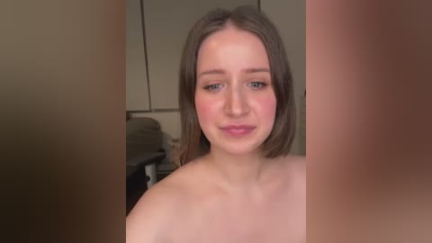 Video of a young, fair-skinned woman with straight, shoulder-length brown hair and light makeup, looking directly at the camera. She is topless, with a blurred, beige kitchen background.