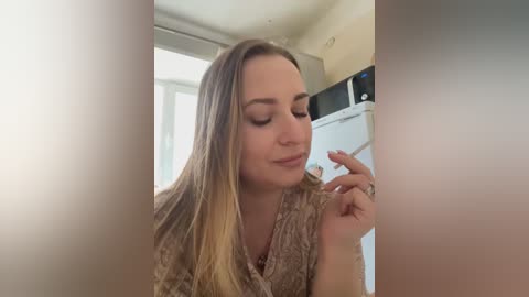 Media: Video of a light-skinned woman with long, straight, blonde hair, wearing a patterned, beige blouse, smoking a cigarette indoors, next to a white fridge with a microwave on top.