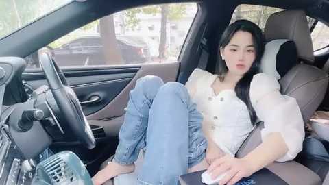 Media: A video of an Asian woman with long black hair, wearing a white blouse and jeans, sitting relaxed in a car with a steering wheel and blurred city background.