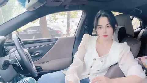 Media: Video of a young East Asian woman with long black hair in a white blouse and blue jeans, sitting in a modern car with beige seats.