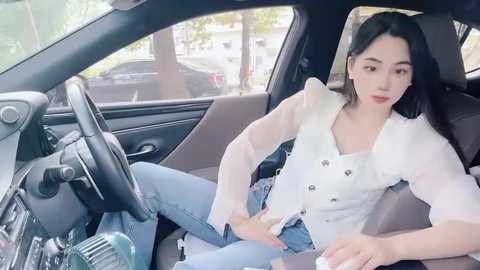 Media: Video of a young East Asian woman with long black hair, wearing a white blouse and blue jeans, sitting in a car with a dashboard view.