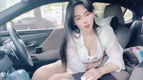 Media: Video of an Asian woman with long black hair, wearing a white blouse unbuttoned to reveal cleavage, sitting in a car with a casual, relaxed expression.