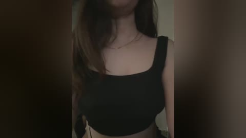 Media: Video of a young woman with long, wavy brown hair, wearing a black, square-neck crop top, indoors with dim lighting.