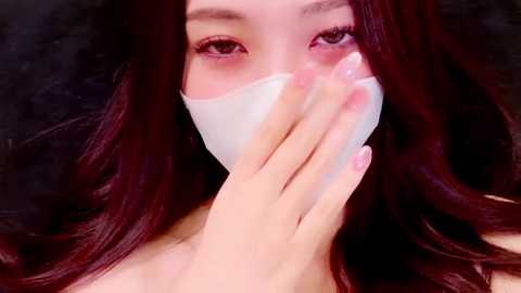 Media: Video of an Asian woman with long, wavy, dark red hair, wearing a white face mask, and covering her mouth with her hand, set against a dark background.