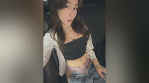 Media: A video of a young woman with fair skin and long brown hair, wearing a black bandeau top, pink lace underwear, and denim shorts, seated in a dark room.