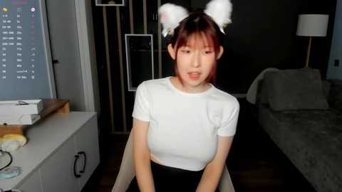 Media: Video of an Asian woman in a white t-shirt, black skirt, and cat ears, sitting in a dimly lit living room with a TV, couch, and dark wooden furniture.