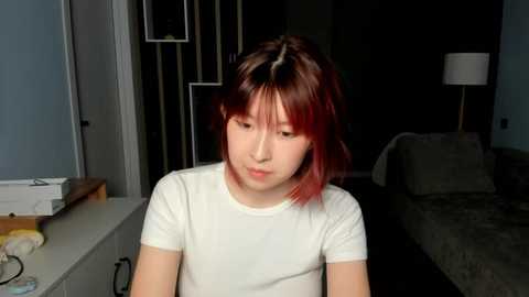 Media: Video of a young Asian woman with short, straight red hair, wearing a white t-shirt, sitting on a white counter in a dimly lit room with a bed and lamp in the background.