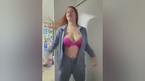 Video of a curvy, red-haired woman in a pink bra and grey hoodie, standing in a dimly lit room with a white wall and colorful posters.